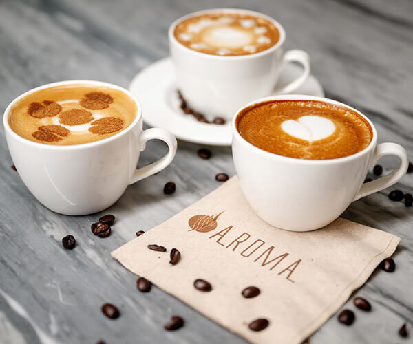 Three coffee cups with different latte art designs on a brown paper napkin with Aroma branding and scattered coffee beans