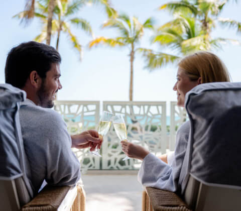 Couple clinking glasses in a cheers with spa robes and a view of the ocean on a sunny day
