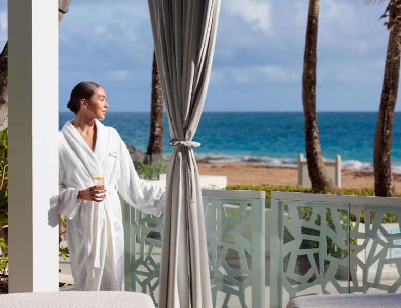 Woman in bathrobe with water in hand looking at ocean view from the spa